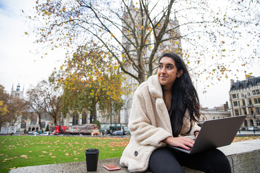 Indian girl works outdoors and uses her laptop, she drinks coffee and she is in a park. Attractive happy female uses technology. Joyful millennial tech woman lifestyle concept