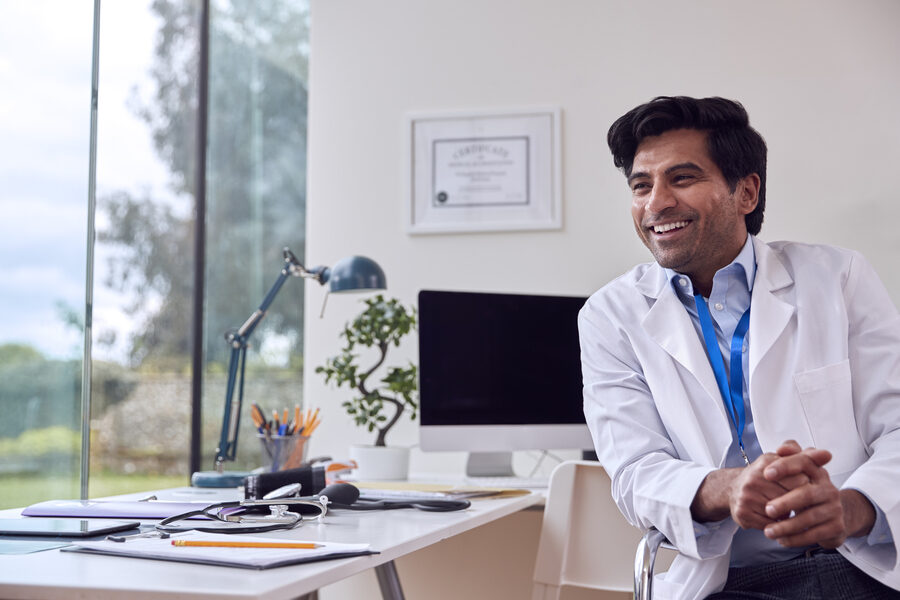 Portrait Of Male Doctor Or GP Wearing White Coat At Desk In Office